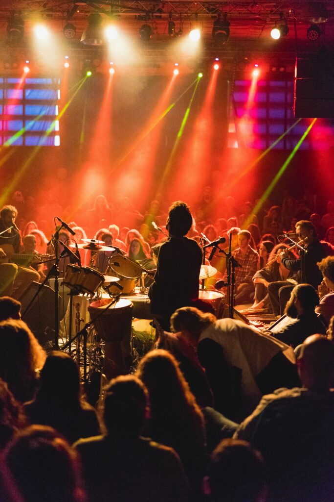 Drummer performing on stage in front of a seated concert audience under colourful lights