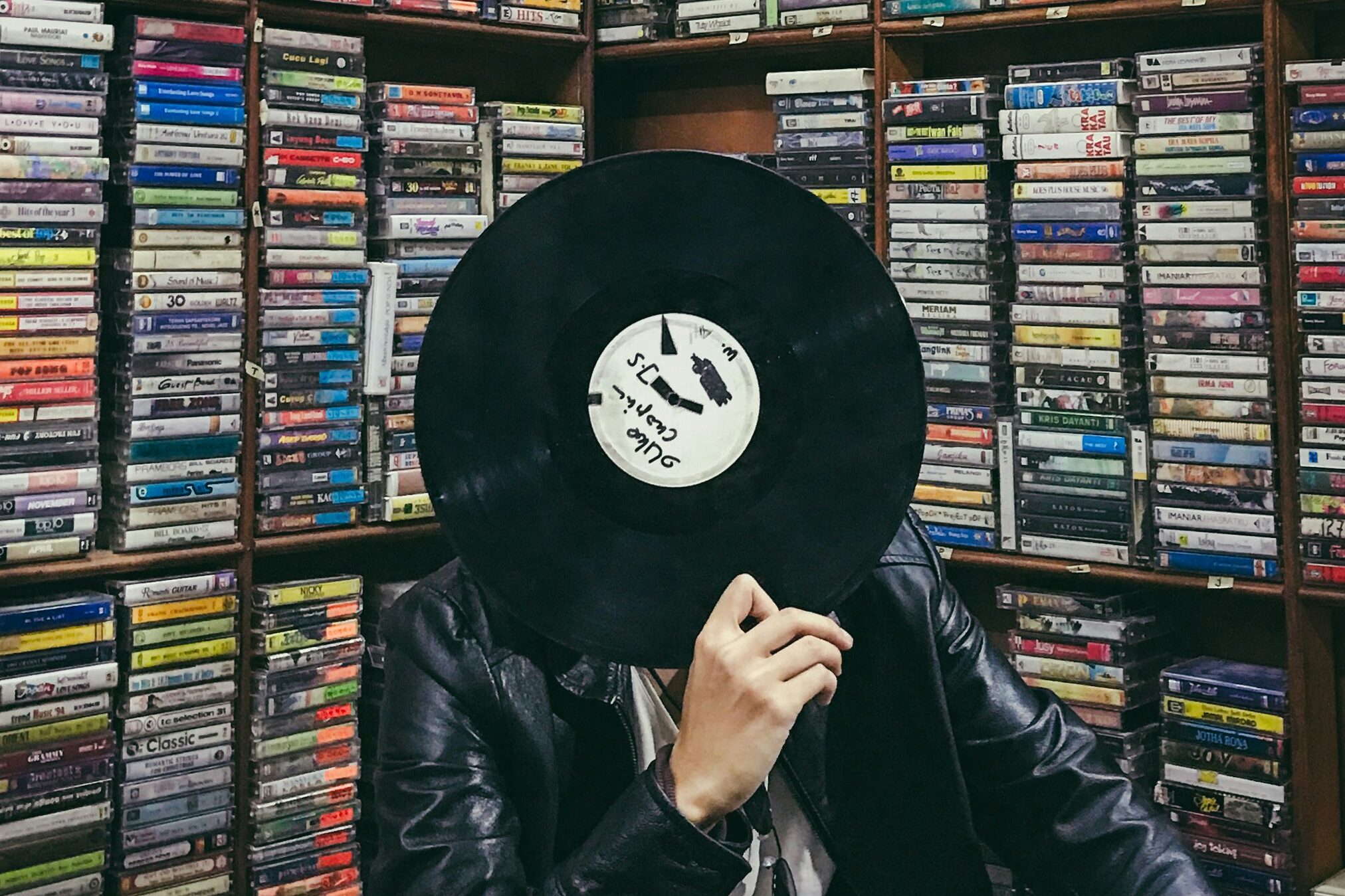 Person holding a vinyl record in front of shelves filled with music cassettes